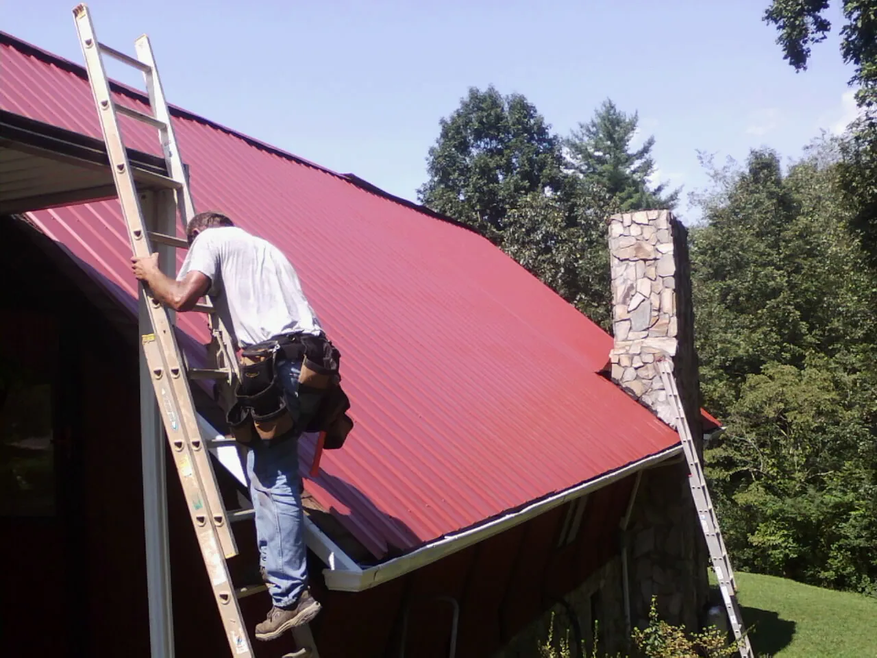 Roofer on a ladder beside a red metal roof and stone chimney — Soffit & Fascia Repair by Roofing Brothers in Hermiston
